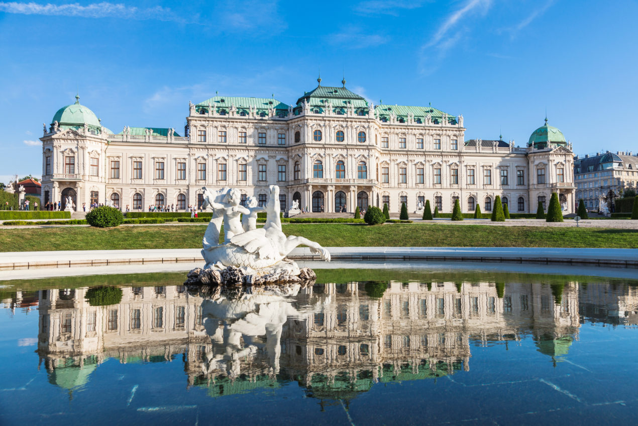 The Belvedere Palace in Vienna, with its ornate Baroque architecture and green domed roofs, reflecting in a large pond. A white statue of nymphs and sea creatures stands in the center of the water.