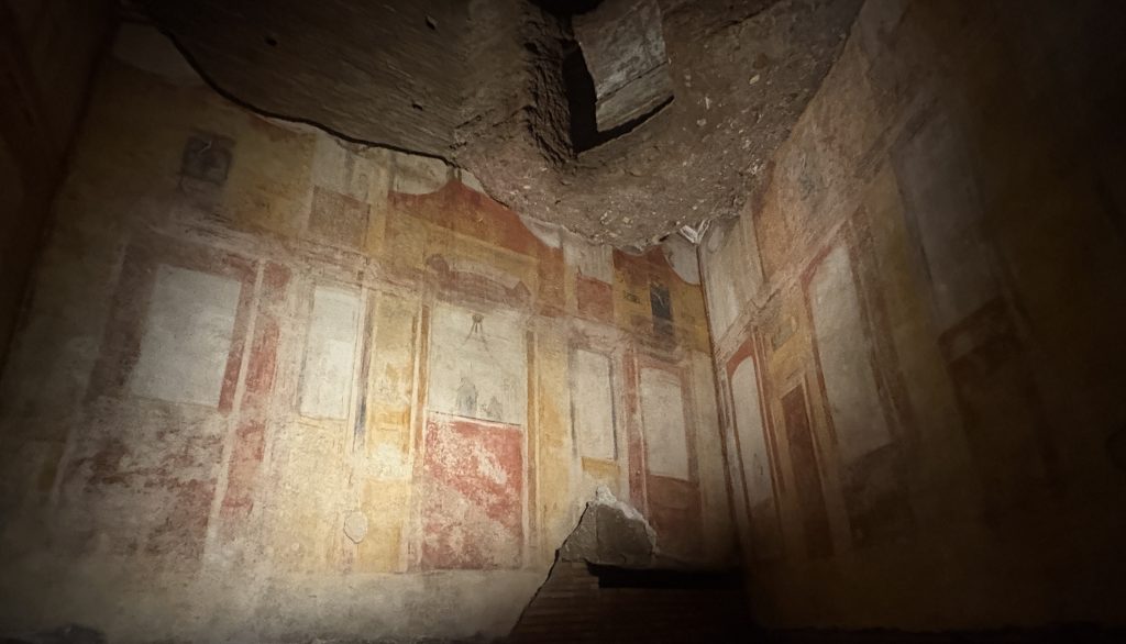 Wall paintings showing architecture in reds and ochres in a dimly lit space underground at the Domus Aurea in Rome, once Nero's Palace.