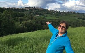 Carol Cram with her arms wide standing in a field in front of the skyline of San Gimignano in Tuscany