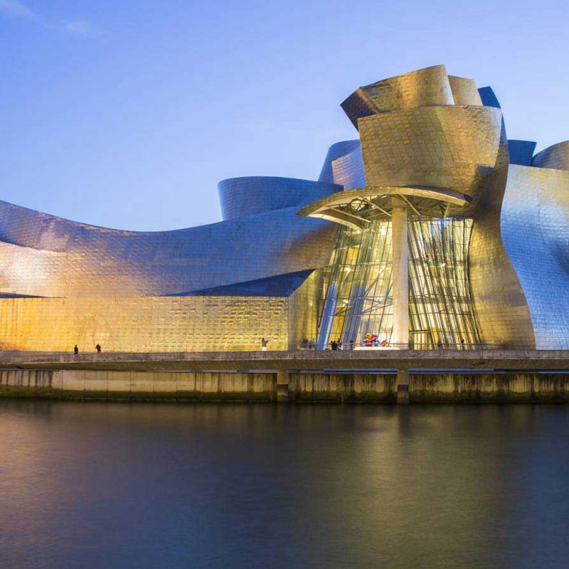The illuminated exterior of the Guggenheim Museum Bilbao at dusk. The modern metallic structure reflects warm lights, with the river running in the foreground.
