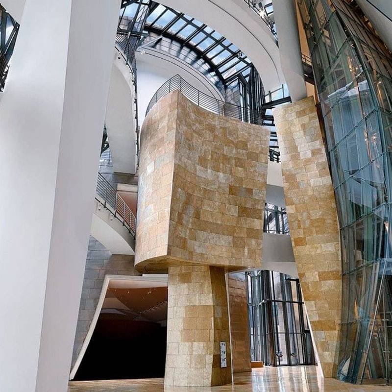 Interior of the Guggenheim Museum Bilbao, showing sweeping architectural curves and stone walls. The modern design features glass and steel elements with natural light pouring through the ceiling.