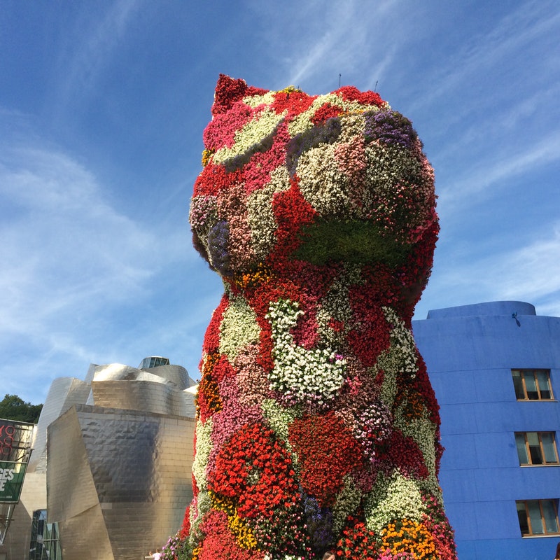 A massive floral sculpture of a dog titled Puppy by Jeff Koons outside the Guggenheim Museum Bilbao. The figure is covered in a vibrant patchwork of colorful flowers against a blue sky.