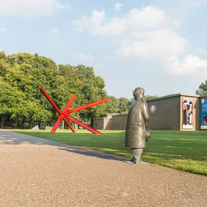 A large red abstract sculpture and a bronze statue of a man stand in front of the Kröller-Müller Museum, set against green trees and a bright sky.