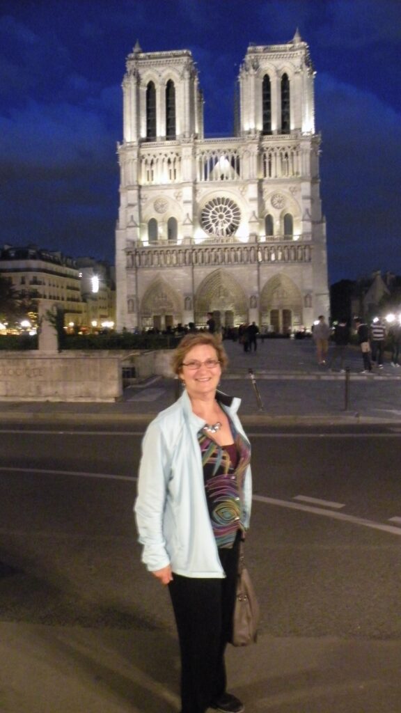 The Artsy Traveler, Carol Cram, in front of a floodlit Notre Dame Cathedral at night in beautiful Paris, France
