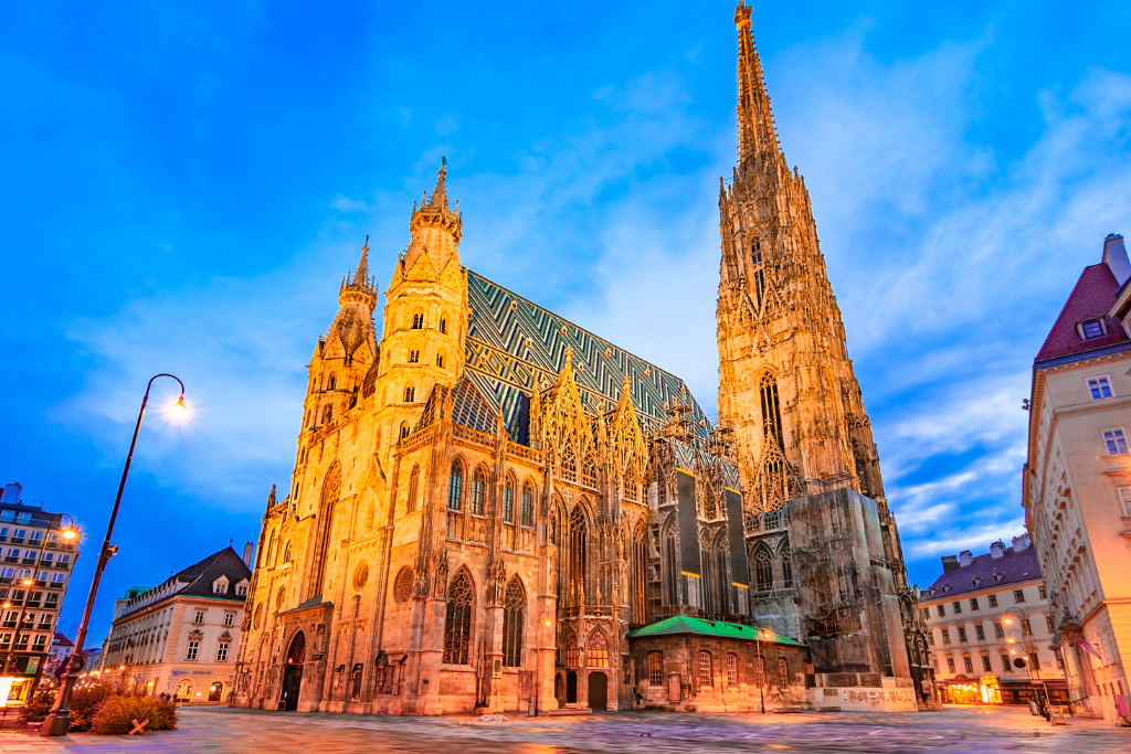 Saint Stephen’s Cathedral in Vienna illuminated at twilight, with its Gothic spires, intricate tiled roof, and ornate facade standing against a deep blue sky. The square around the cathedral is lit by street lamps.