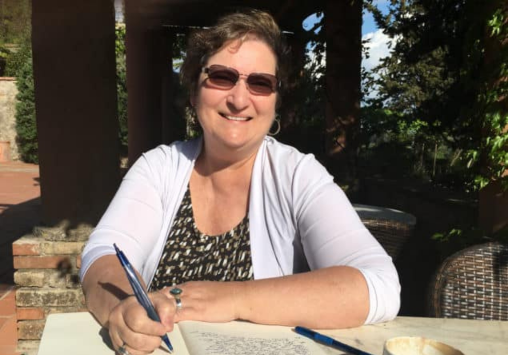 The Artsy Traveler, Carol M. Cram, working on one of her arts-inspired novels at an outdoor table in Tuscany.