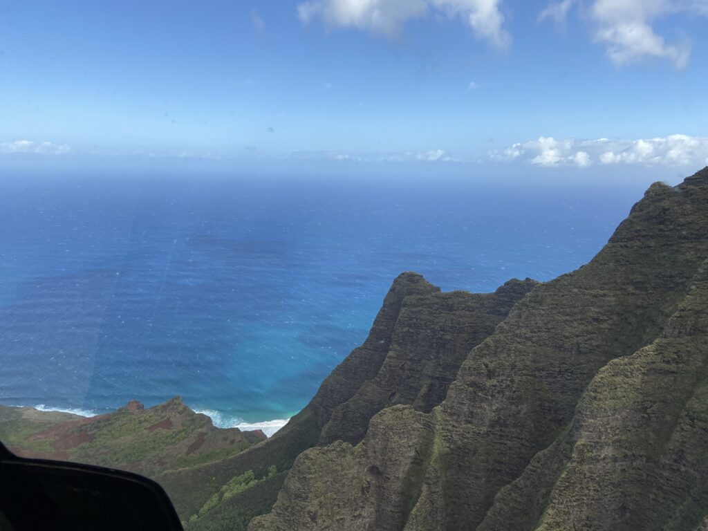 View of deep blue water with sharp green cliffs in the foreground seen from a helicopter ride over Kauai