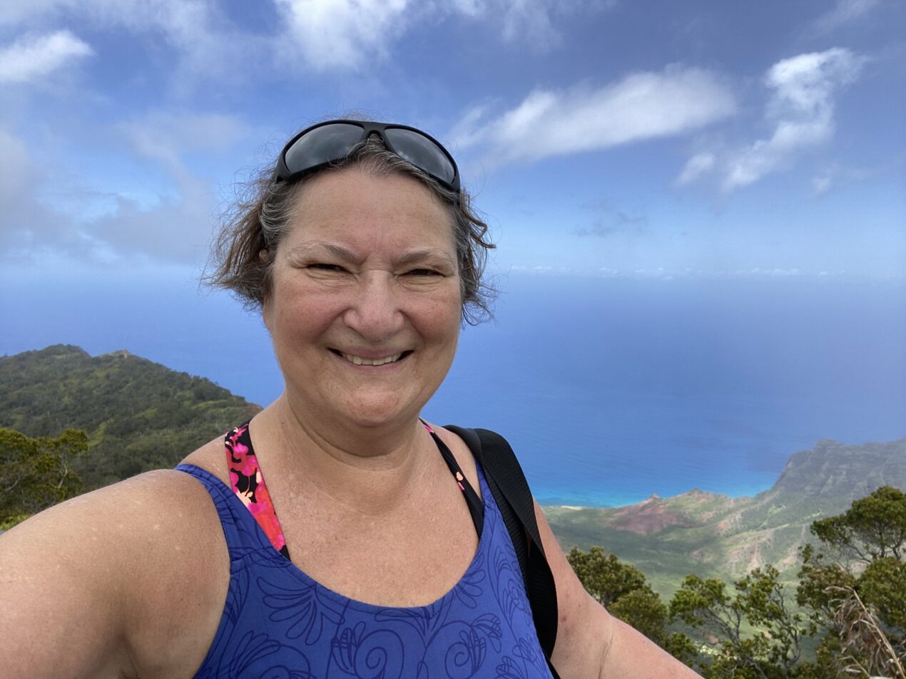 Carol Cram taking a selfie in front of a breathtaking vista of green hills, blue ocean, and scattered clouds. They are wearing a blue patterned tank top and sunglasses resting on their head.