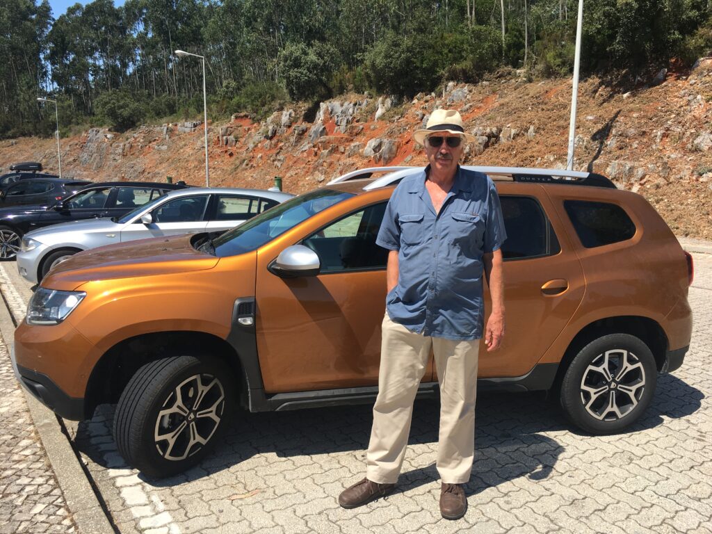 Gregg Simpson standing in front of an orange rental car in Portugal. Driving is not green travel unfortunately.