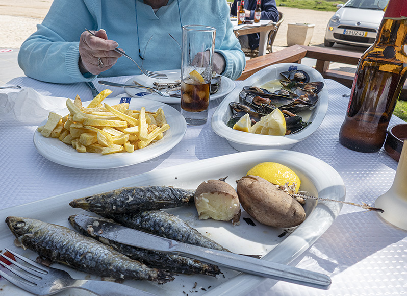 Outdoor table with food - french fries, whole fish, mussels, bottle of beer. Traveling to Europe in the age of COVID means lots of outdoor meals.