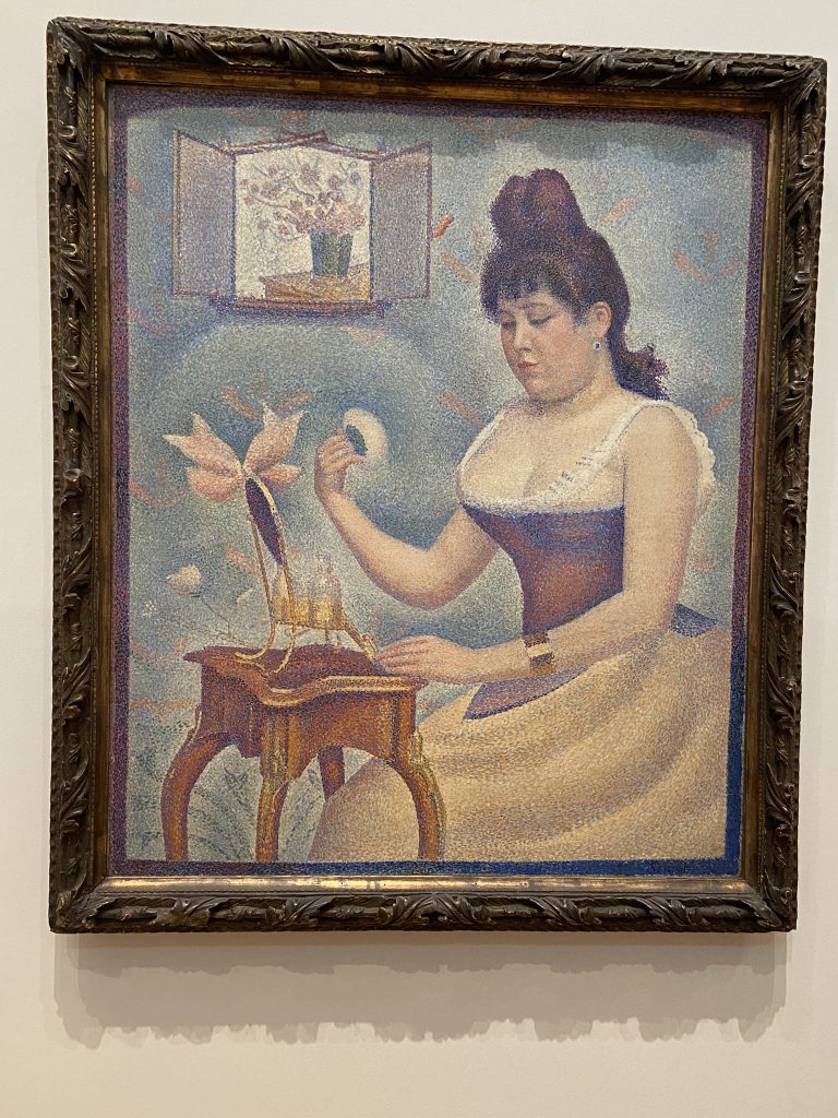 Painting called "Young Woman Powdering Herself" by Goerges Seurat showing a woman wearing a red corset and holding a powder puff. The style is pointillist and the piece is included in the Impressionist collection at the Courtauld Gallery in London.