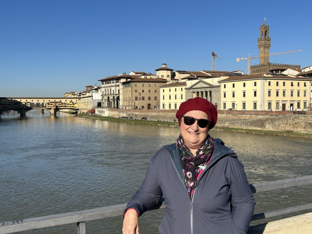 Carol Cram on a bridge across the Arno with the Palazzo Vecchio and Ponte Vecchio in the background in Florence.