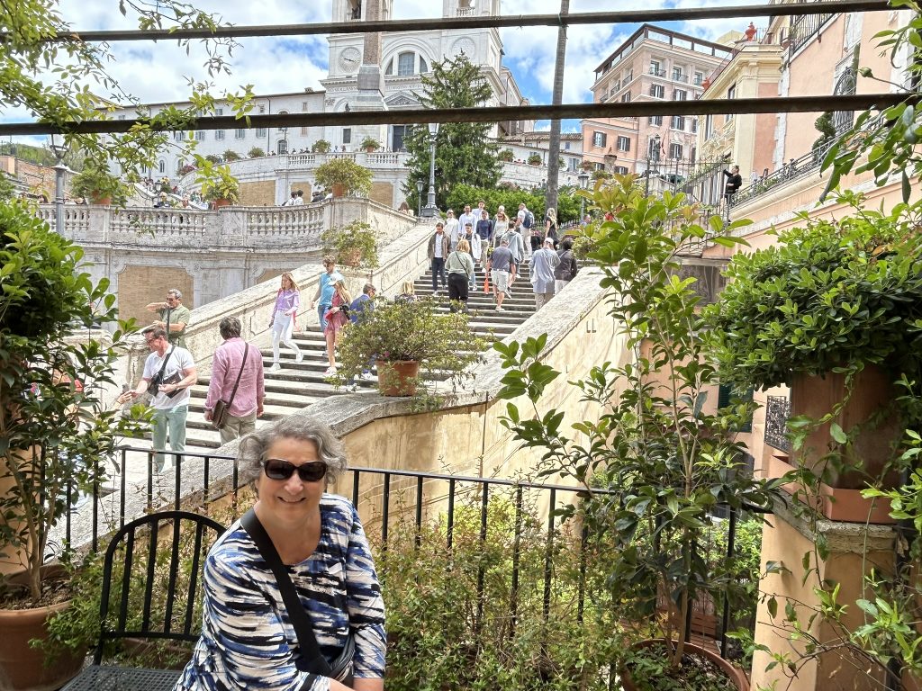 A vibrant view of the Spanish Steps in Rome, adorned with colorful flowers, leading up to the twin towers of Trinità dei Monti Church. Visitors gather around the piazza in front of the steps.