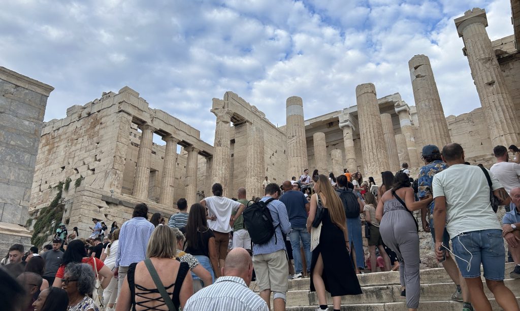 Massive columns at the entrance to the Acropolis in Athens visible as you climb up to the Acropolis. the backs of a large crowd of people climbing to the top is in the foreground.