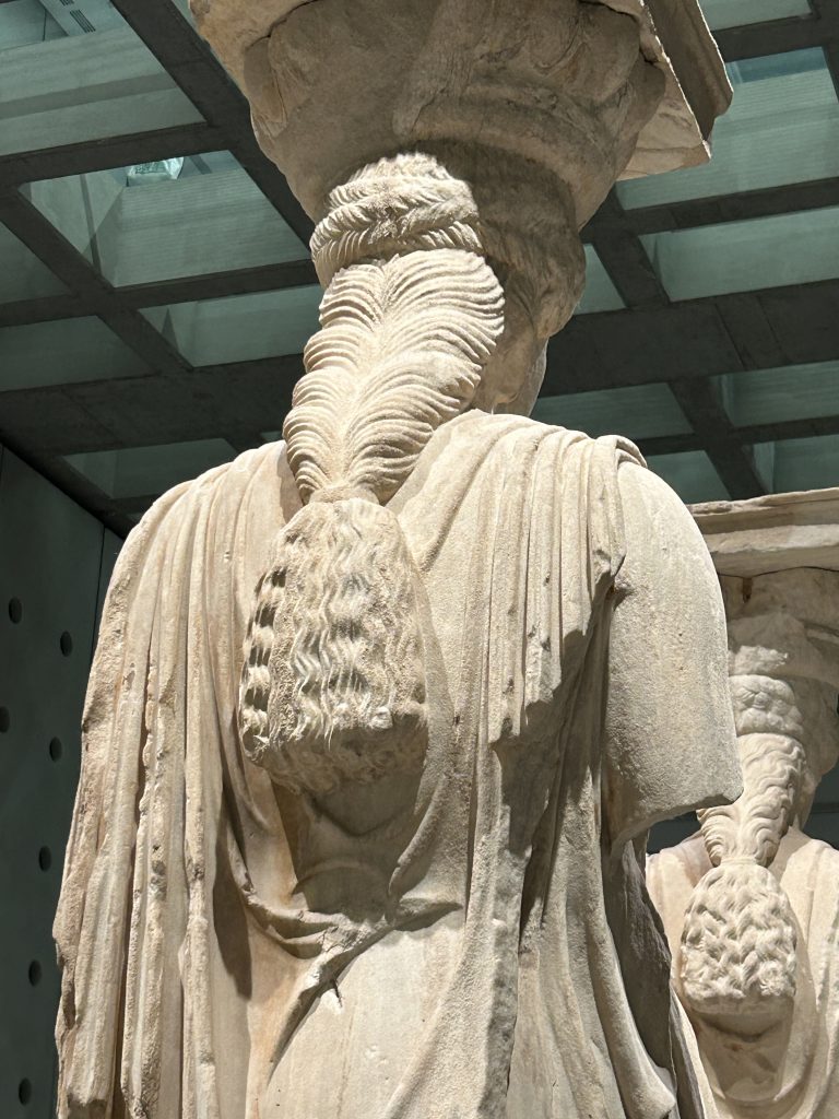 Close-up of the back of one of the female statues wearing Greek robes with a pediment on top of her head. The view shows the statue's intricately braided hair. This is one of the original columns used to hold up the Erechtheion Temple on the Acropolis, and is very classically Greek and beautiful. The column is displayed in the Acropolis Museum--a must-see museum in Athens.