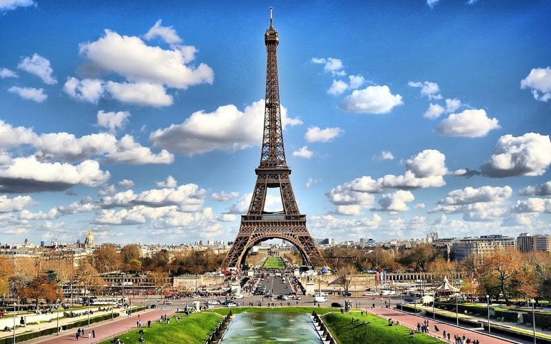 A classic view of the Eiffel Tower taken from the Trocadéro gardens, with fountains, greenery, and a bright blue sky with fluffy clouds.