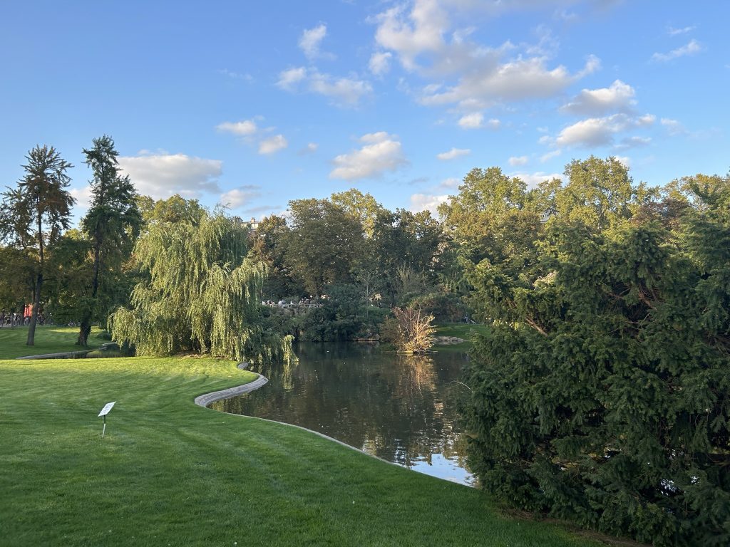 A serene view of a lush garden with a pond surrounded by trees and bushes, under a bright blue sky with scattered clouds.