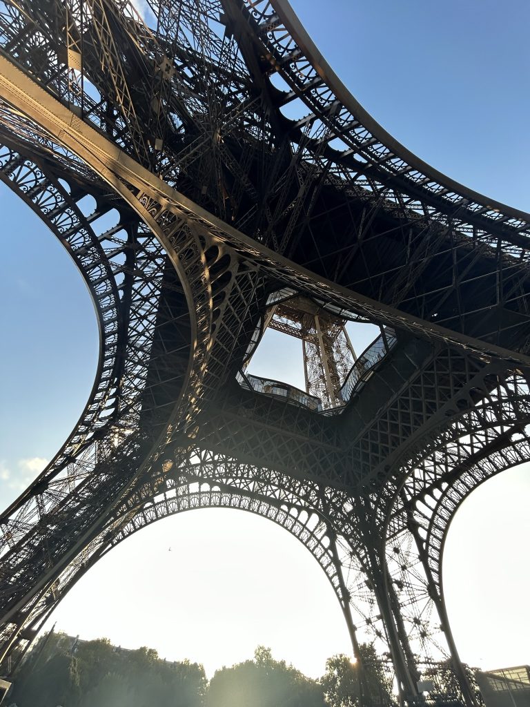 View from below the Eiffel Tower showcasing its intricate iron latticework structure against a bright blue sky, with sunlight streaming through the open arches.