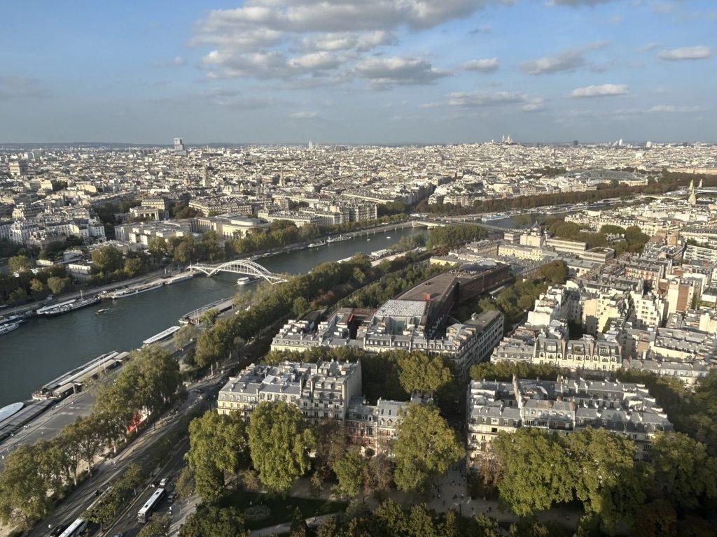 A sweeping view of Paris from above, capturing the Seine River, bridges, and iconic Parisian architecture under a partly cloudy sky.