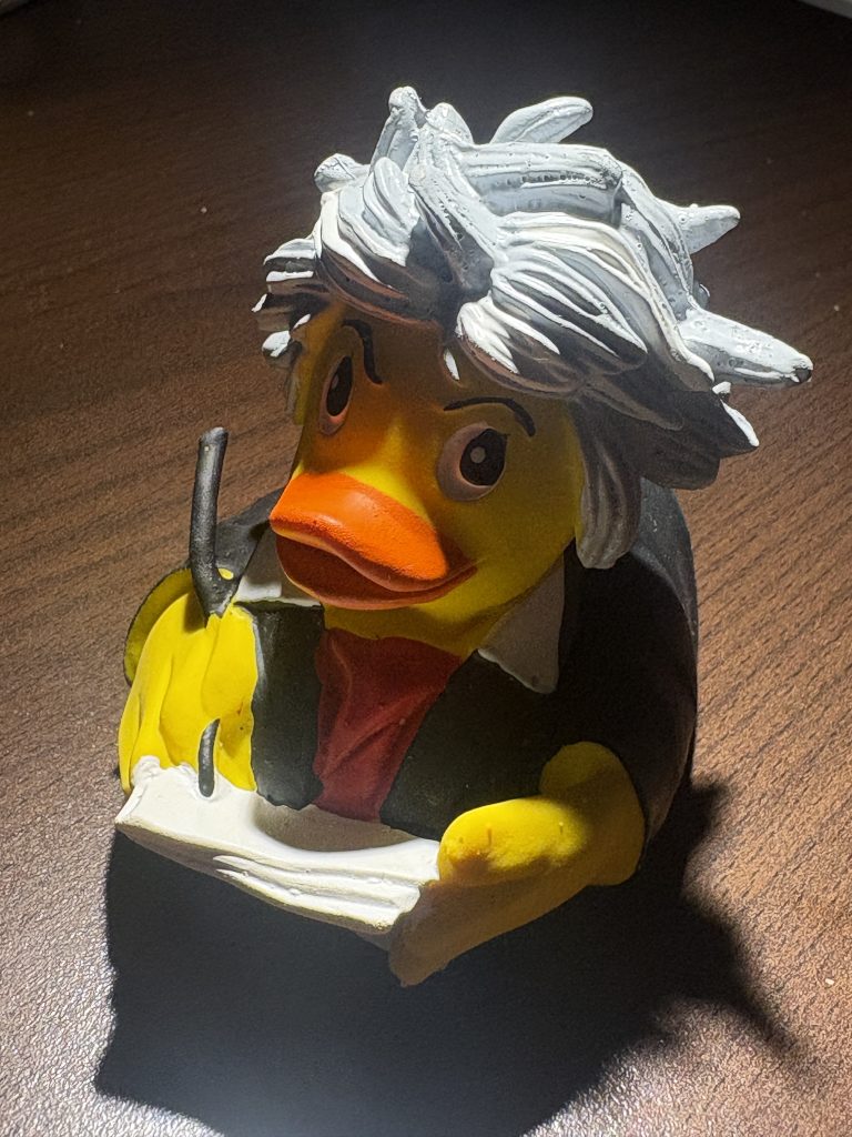 Plastic ruber duckie representing Bewethoven with wild white hair and holding a quill and a pad of paper