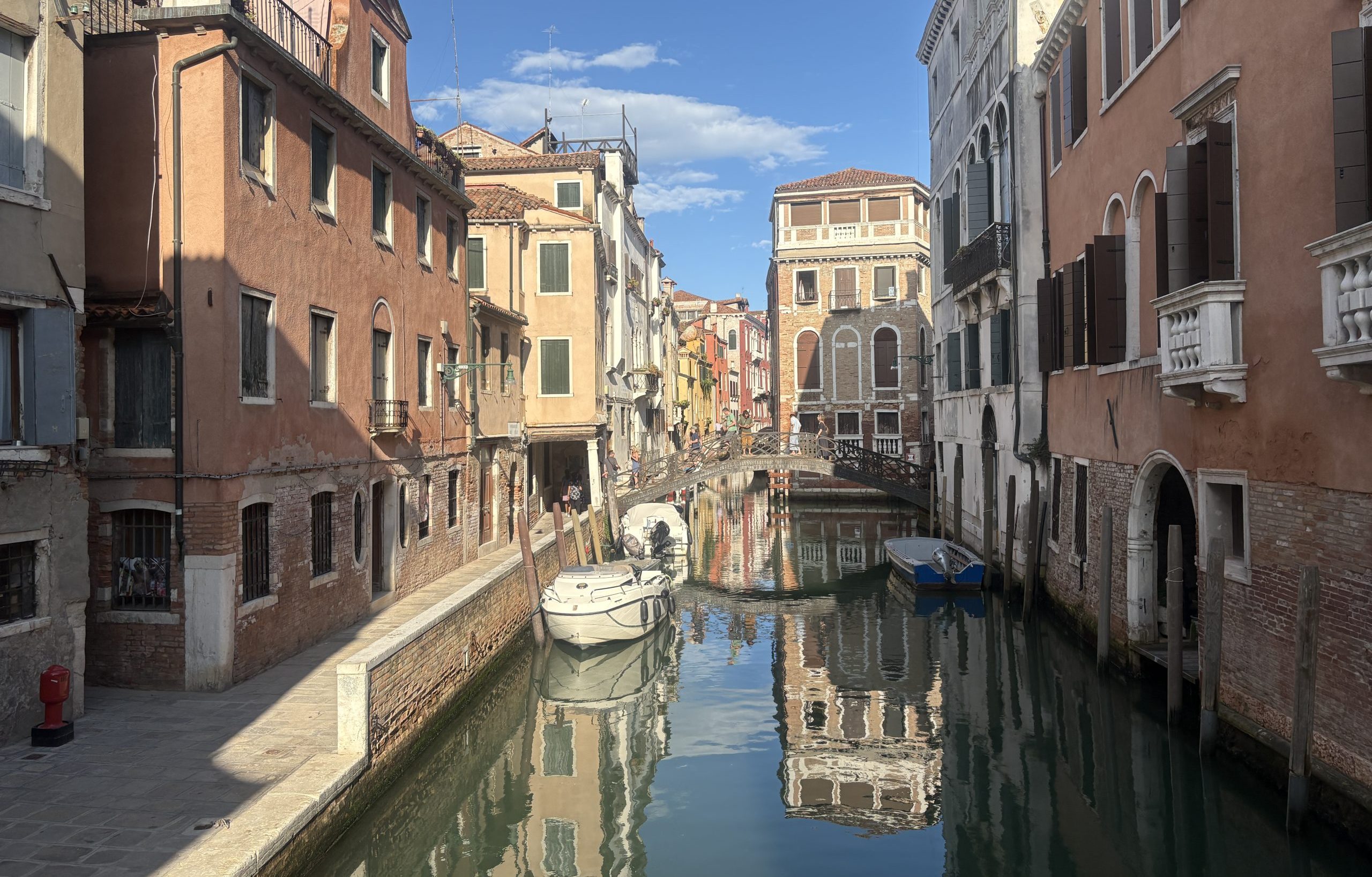 A quiet side canale and street in Venice with buildings and boats reflected in the calm water with a blue sky above.
